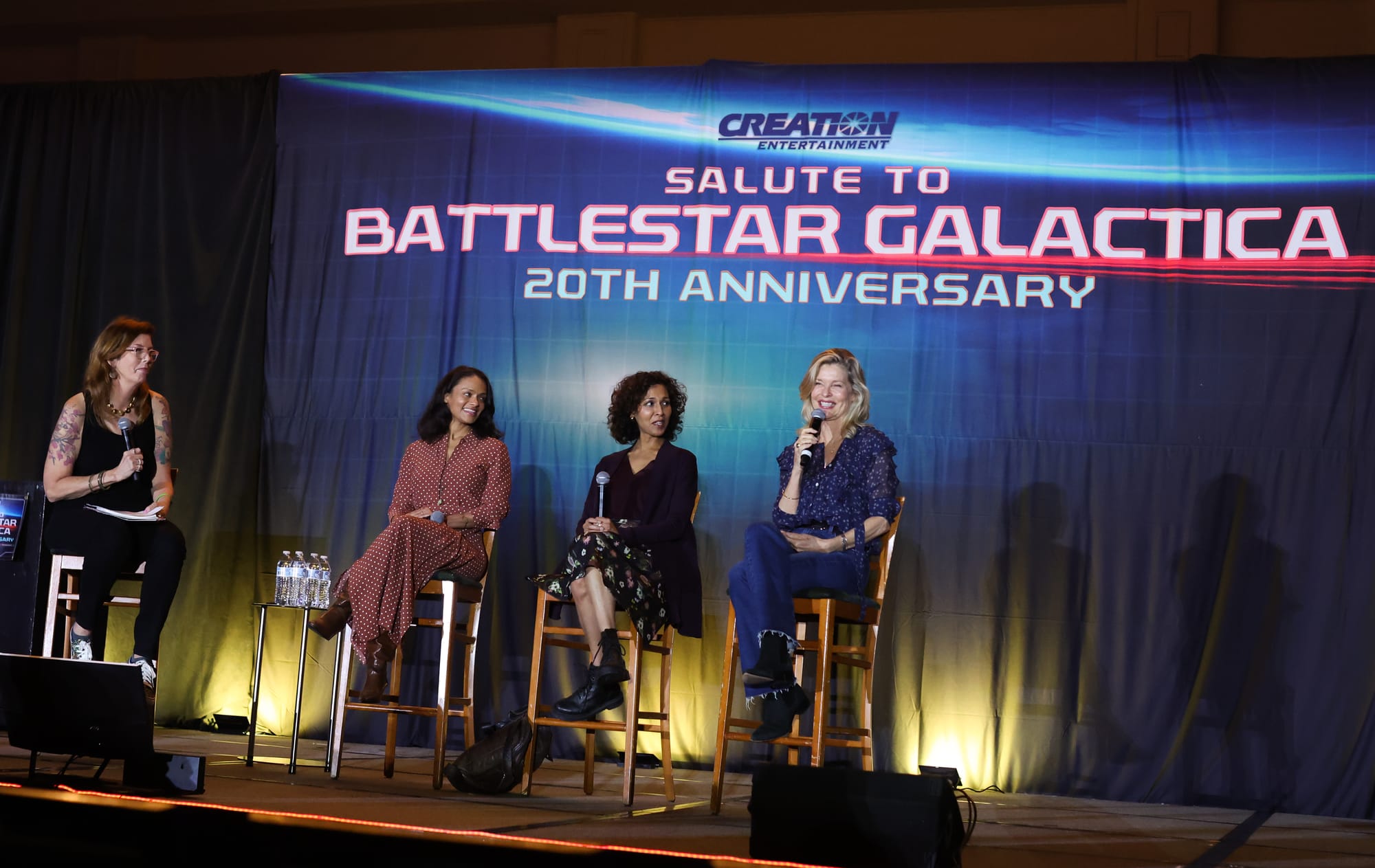 Mo Ryan, Kandyse McClure, Rekha Sharma and Kate Vernon seated on stage against a blue backdrop that says Creation Salute to Battlestar Galactica