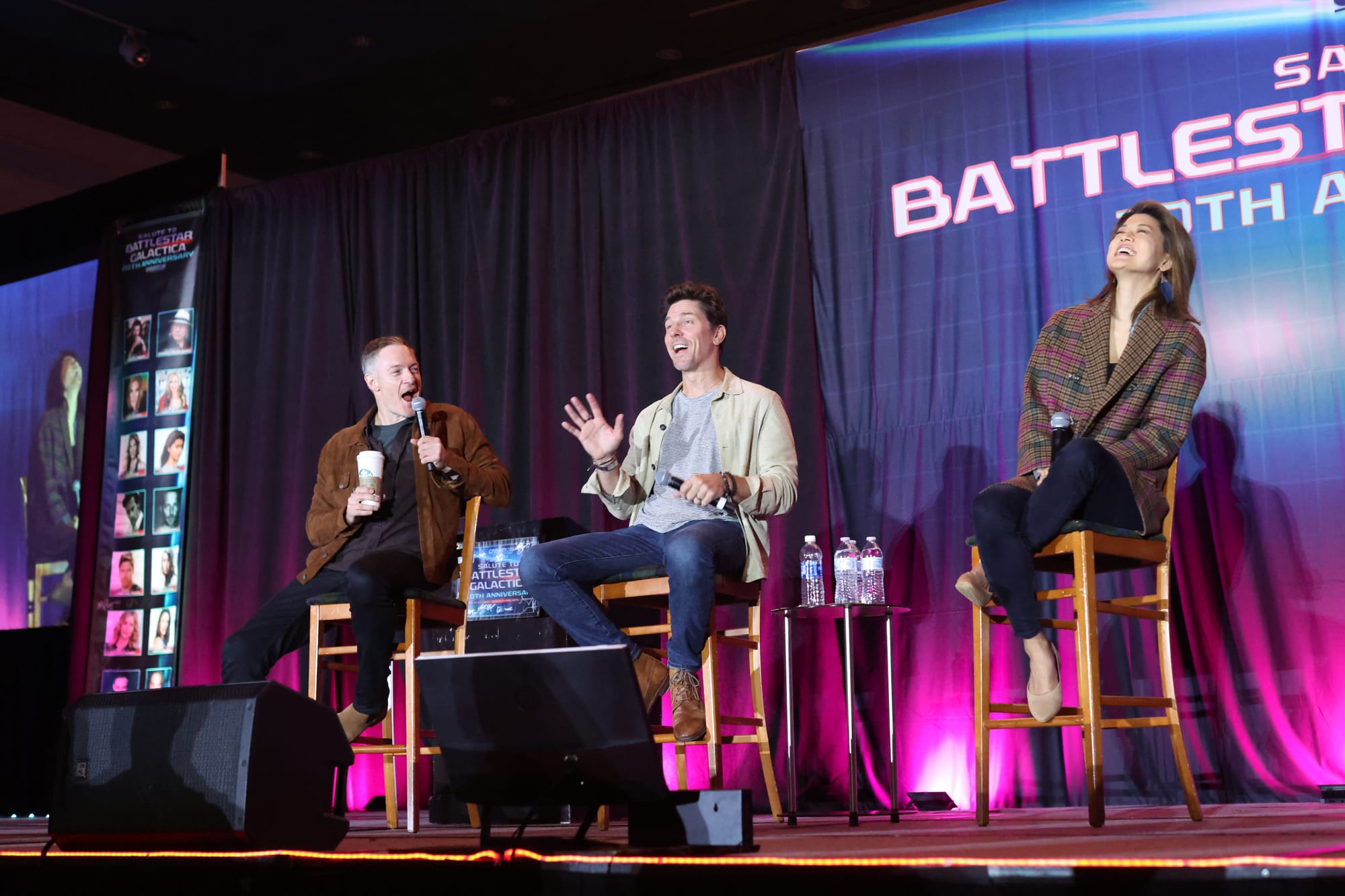 Tahmoh Penikett, Michael Trucco and Grace Park seated on chairs on a stage with a blue and purple backdrop.