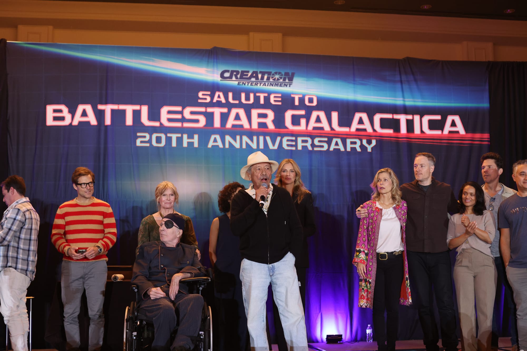 About a dozen actors from Battlestar Galactica stand on a stage as Edward James Olmos speaks