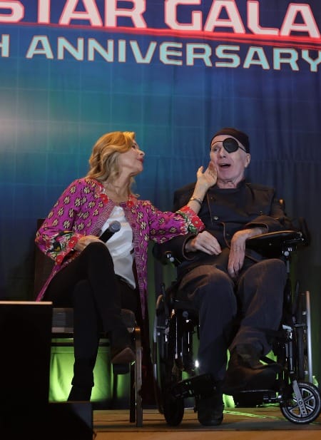 Kate Vernon strokes the cheek of Michael Hogan, as both are seated on a stage with a blue backdrop