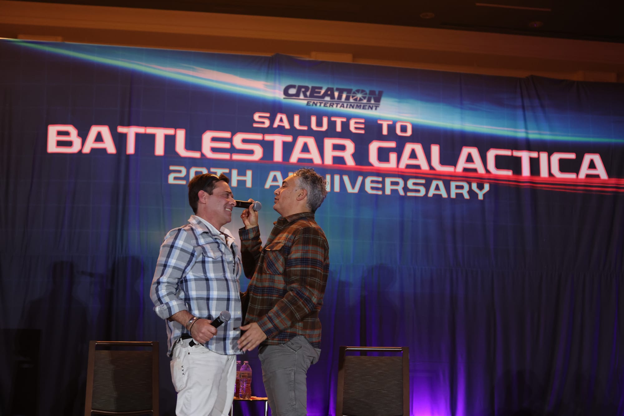 James Callis and Alessandro Juliani stand facing each other on a stage with a blue backdrop and the words Creation Salute to Battlestar Galactica