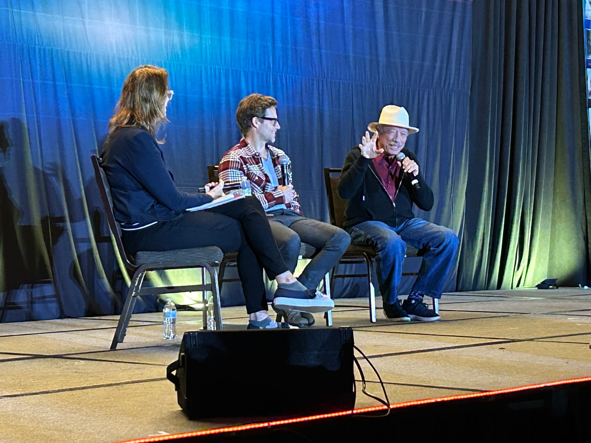 Mo Ryan, Jamie Bamber and Edward James Olmos seated on a stage with a dark blue background. 