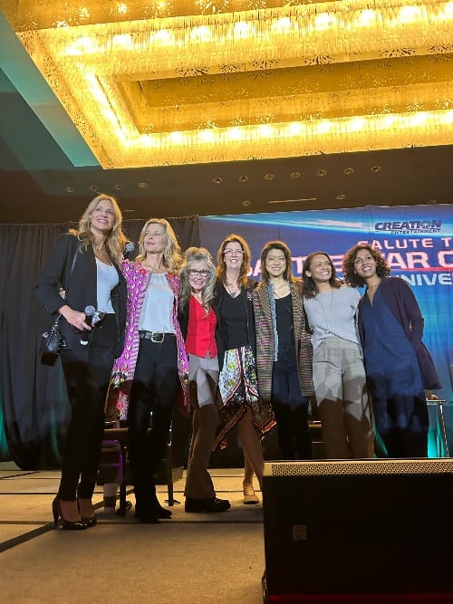 Tricia Helfer, Kate Vernon, Mary McDonnell, Mo Ryan, Grace Park, Kandyse McClure and Rekha Sharma standing on stage with bright lights above