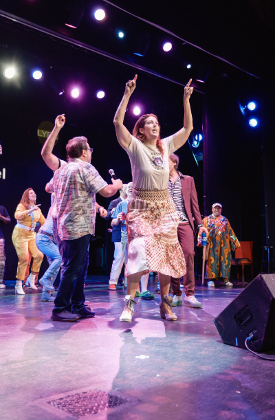 A woman on a stage lifts her hands up at a musical event, with other people in the background. 